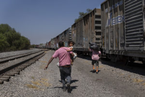 Menschen, mit Gepäck und einem Kind auf dem Arm, rennen hinter einem braunen Güterzug her.