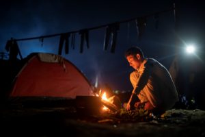 Ein junger Mann sitzt am Feuer ei Nacht, im Hintergrund ein Zelt.